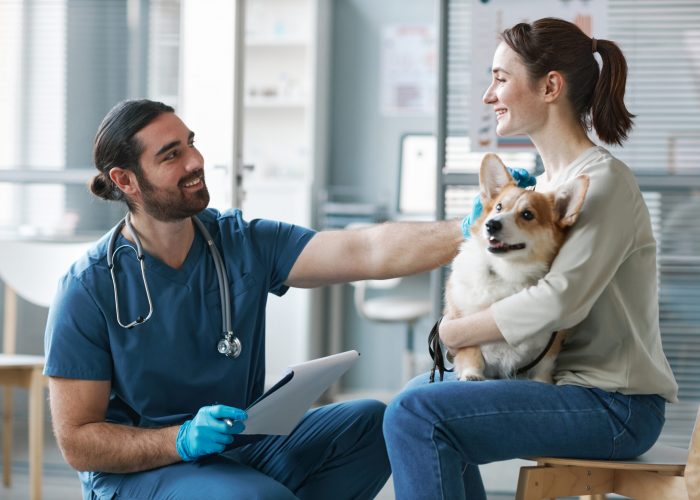 Happy young veterinarian in gloves and uniform consulting female owner of purebred welsh pembroke corgi dog in medical office